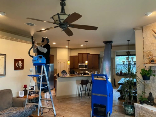 A technician is cleaning an air duct in the ceiling while standing on a ladder.